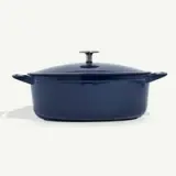 A navy blue, oval-shaped cast iron Dutch oven with a lid and side handles is displayed against a white background.