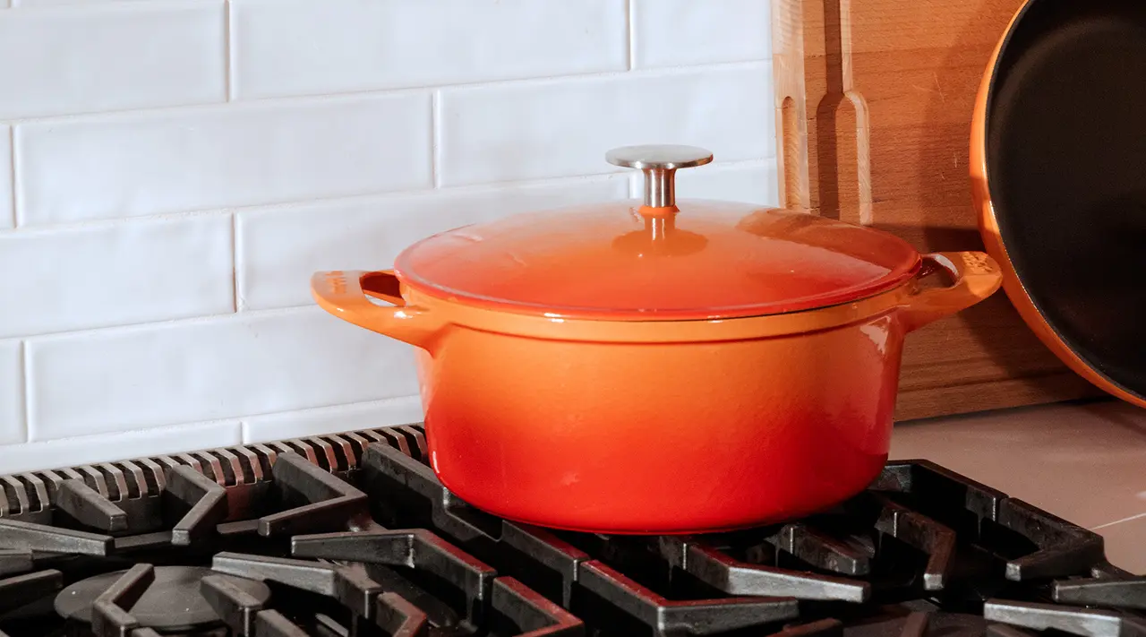 An orange enameled cast iron pot sits atop a stove burner against a backdrop of white subway tiles.