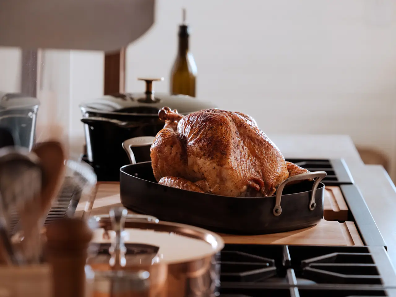 A roasted turkey in a roasting pan on a stove surrounded by cooking utensils, hinting at a holiday or festive meal preparation.