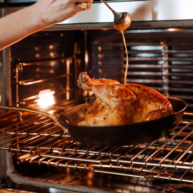 A person is basting a golden-brown roasted chicken fresh from the oven.