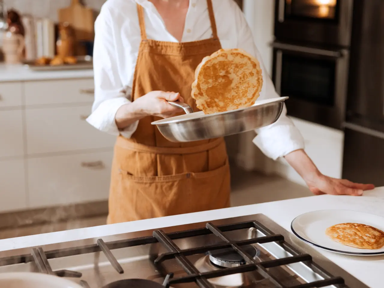 A person in a brown apron is cooking pancakes in a kitchen, flipping one pancake with a pan.