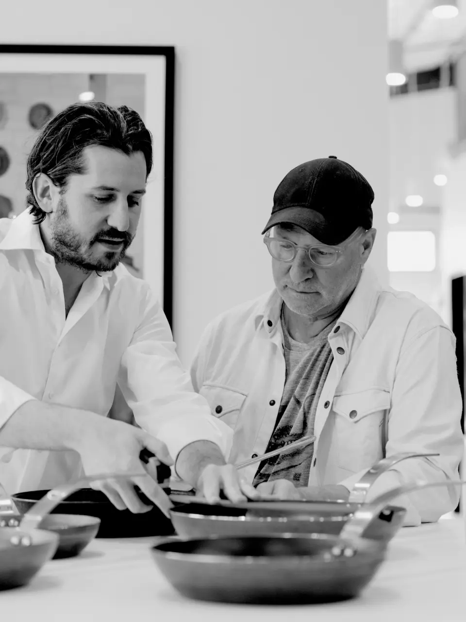 Two men, one younger with a beard and a white shirt, and an older man wearing a cap, are intently focused on something in a pan they are both handling, in a black and white photo.