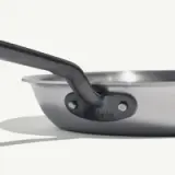 A stainless steel frying pan with a long handle is showcased against a light background, casting a sharp shadow.