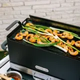 Shrimp and green onions are sizzling on a black grill, with a wooden spoon and sauce in a bowl nearby.