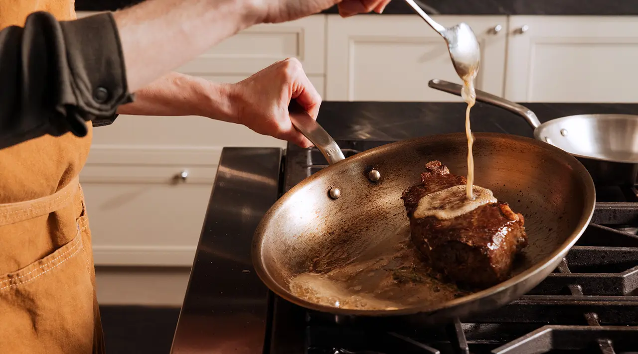 A person in an apron drizzles sauce over a sizzling steak in a pan on the stove.