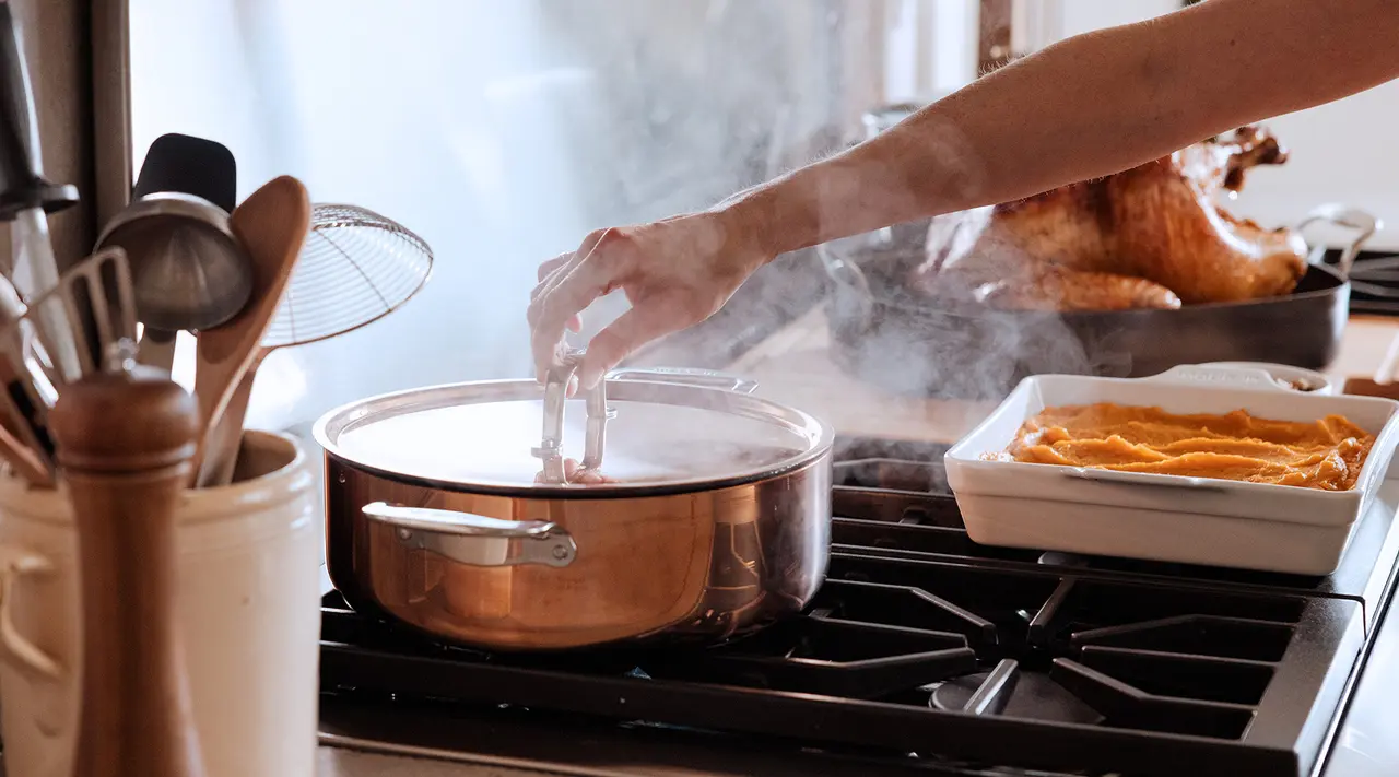 A person is cooking in a kitchen with steam rising from a pot on the stove and a roasted turkey and casserole dish nearby on the countertop.