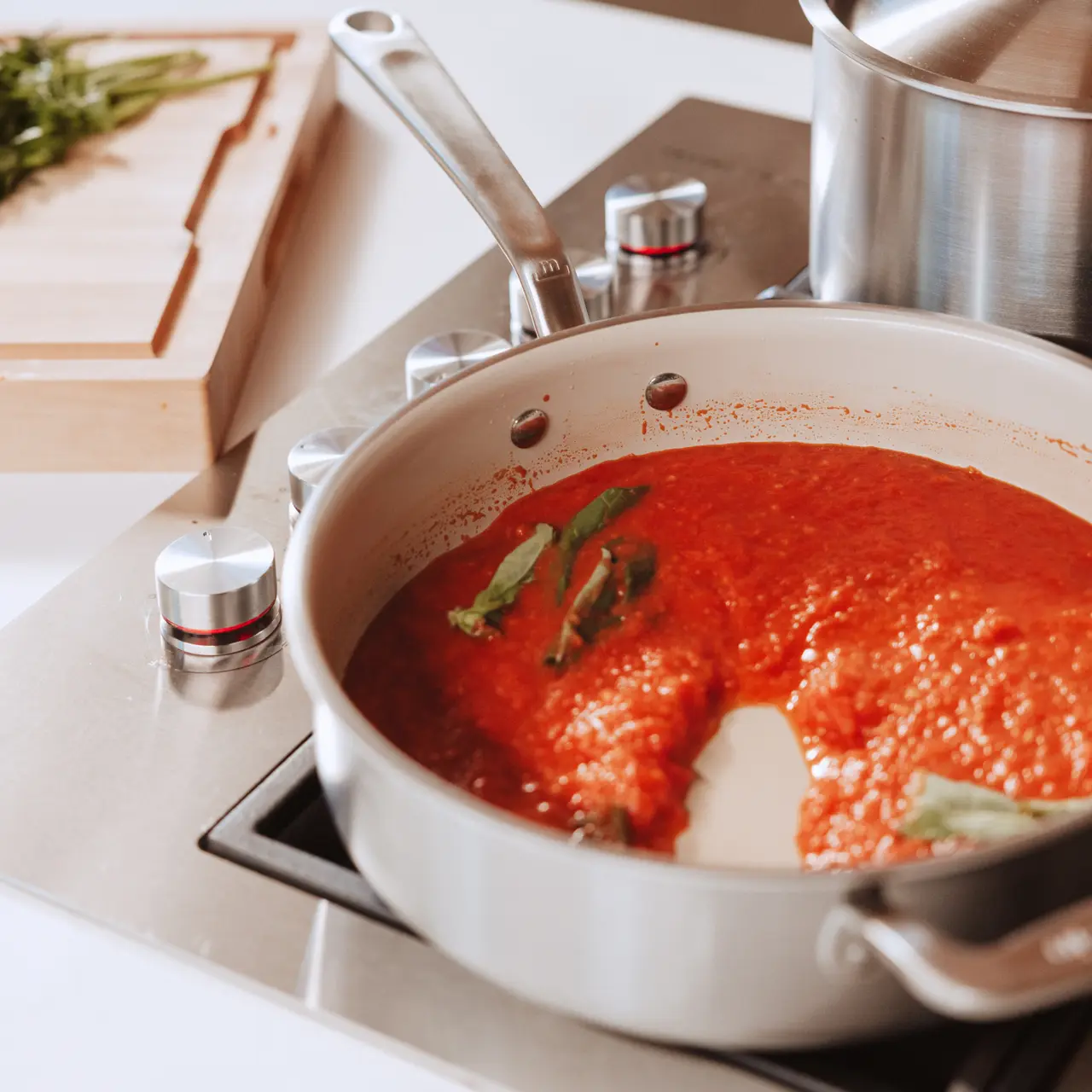 A white pan of red sauce with green herbs sits on a stovetop next to a wooden cutting board.