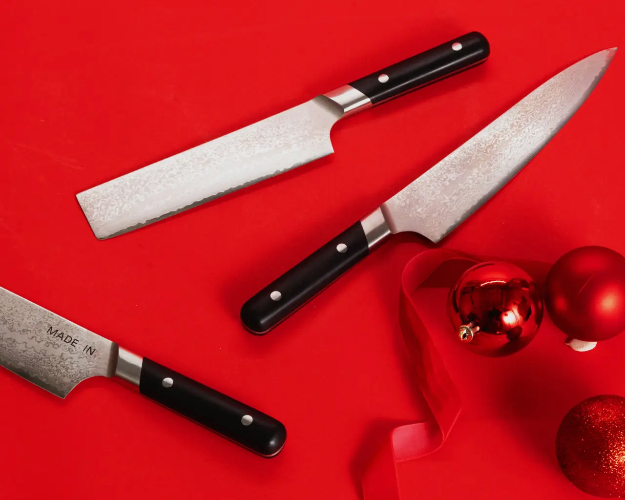 Three kitchen knives with black handles and stainless steel blades are arranged on a festive red background alongside decorative Christmas ornaments.