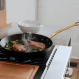 Two steaks are being cooked in a frying pan with vegetables on a kitchen stove, with steam rising from the pan.