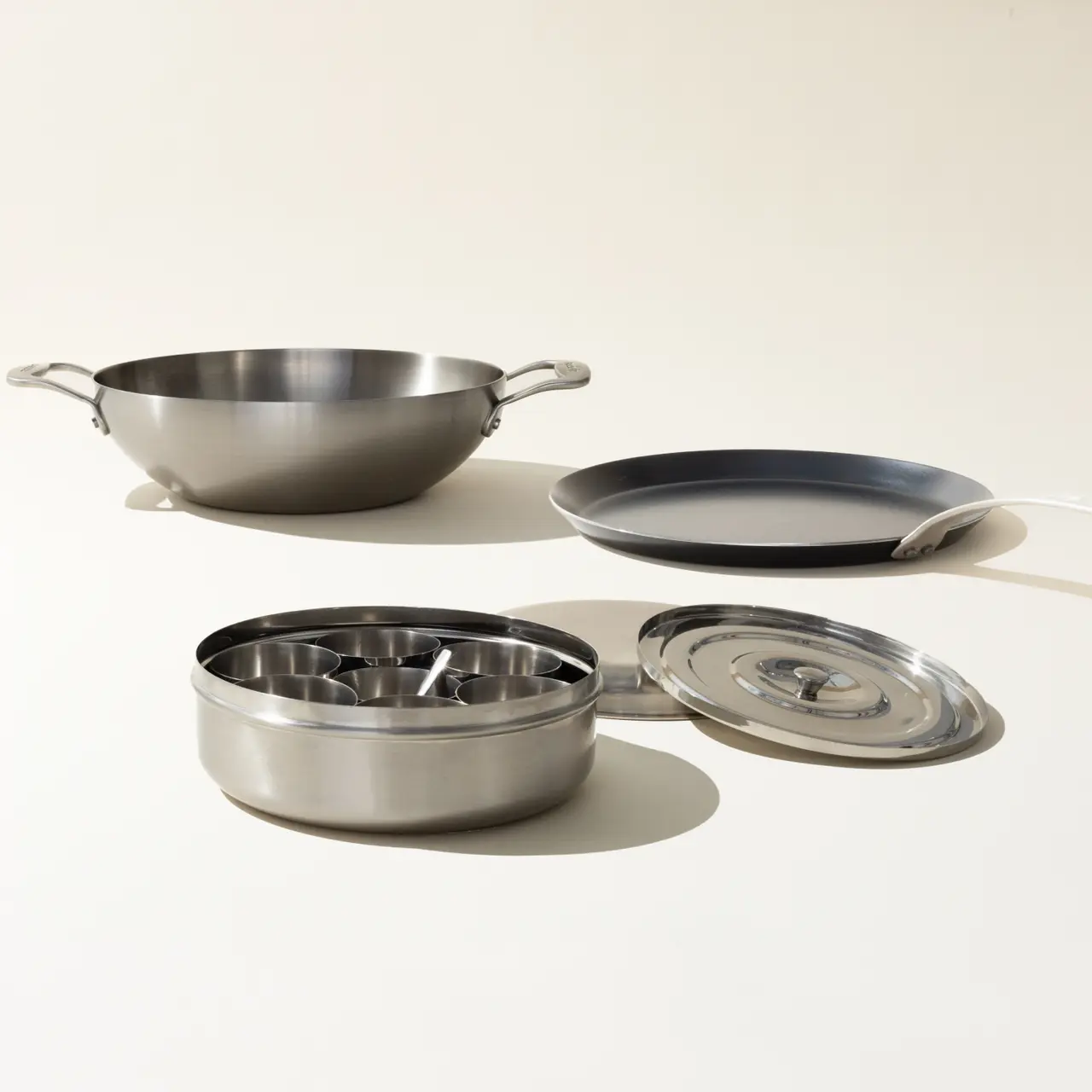 A set of stainless steel cookware, including a wok, skillet, and saucepan with a lid, is arranged on a light background.