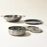 A set of stainless steel cookware, including a wok, skillet, and saucepan with a lid, is arranged on a light background.