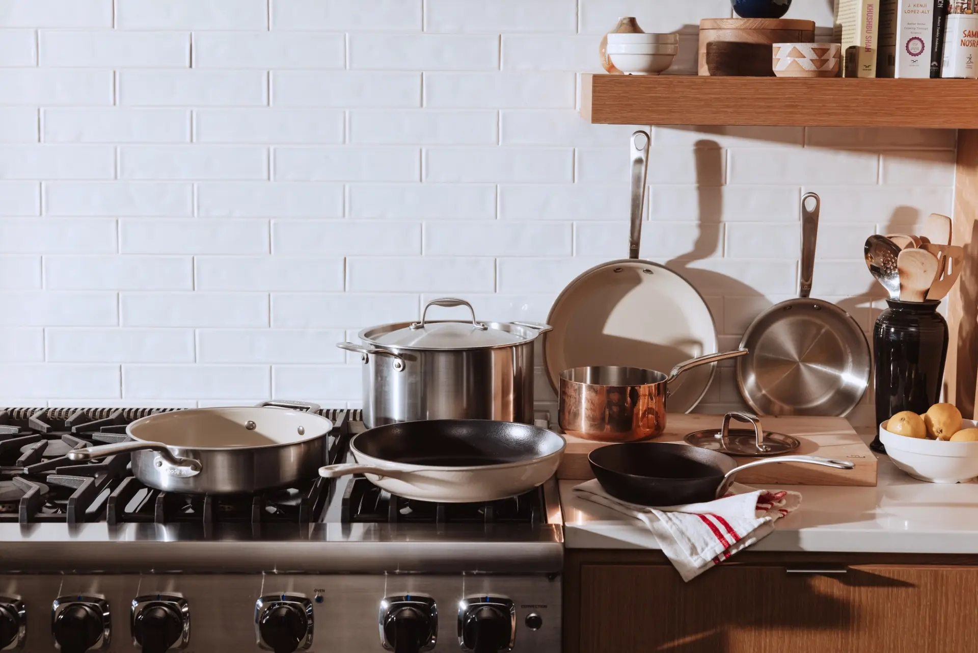 A variety of cookware, including pots and frying pans, is arranged on a kitchen counter beside a stove, with a shelf holding additional kitchen items above.