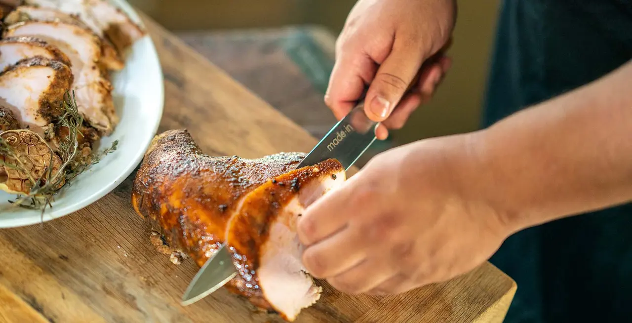 A person is slicing a crispy, roasted pork loin on a wooden cutting board.