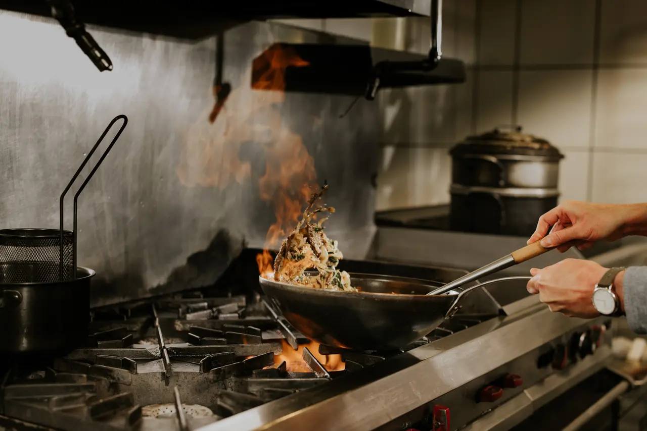A chef is sautéing food in a pan on a stove with flames leaping from the pan, indicating a cooking technique called flambé.
