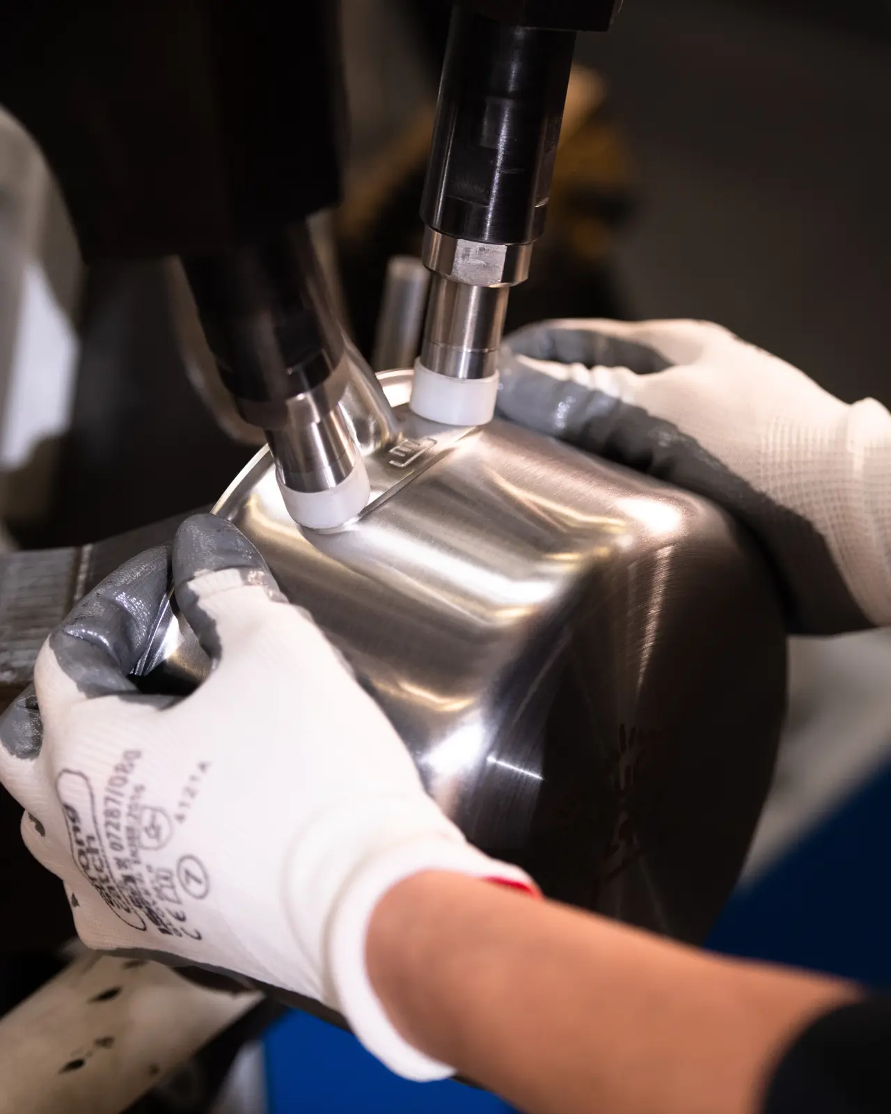 A pair of hands wearing white gloves holds a metallic part while a machine applies a process to its surface.