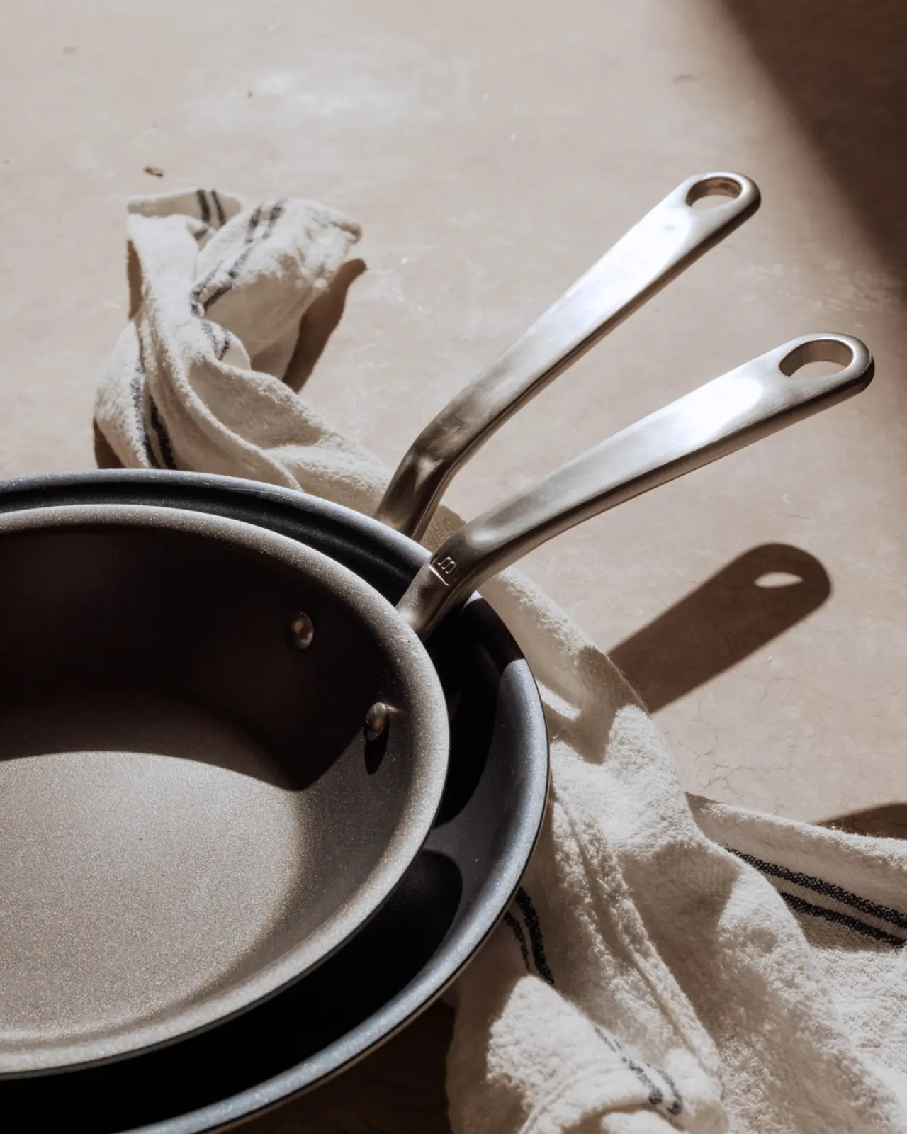 Two frying pans with stainless steel handles rest on a dishtowel on a textured surface, casting soft shadows.