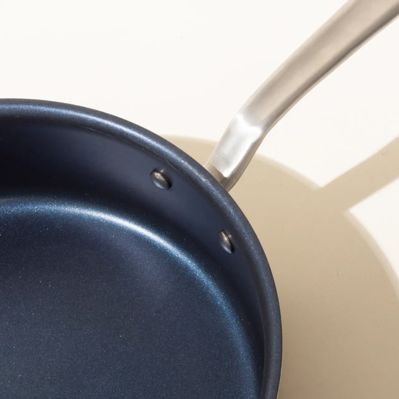 A close-up view of a blue non-stick frying pan with a silver handle, highlighting the attachment point of the handle to the pan.