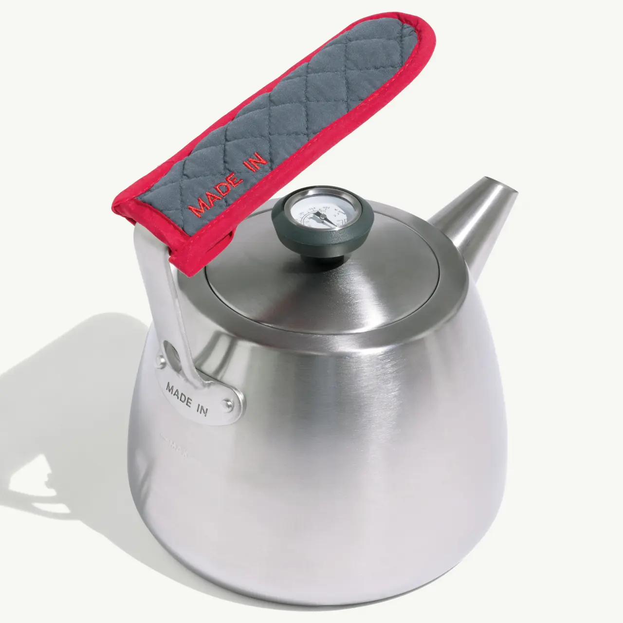 A stainless steel kettle with a gray and red quilted handle and a lid featuring a small thermometer.