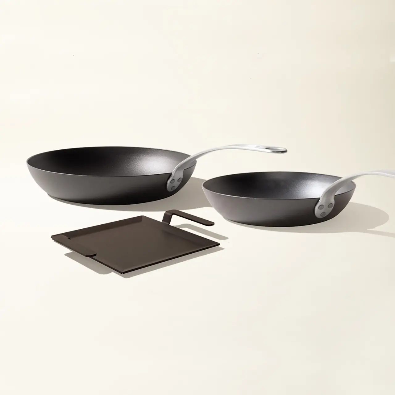 Two black frying pans with silver handles are accompanied by square, dark trivets against a light background.