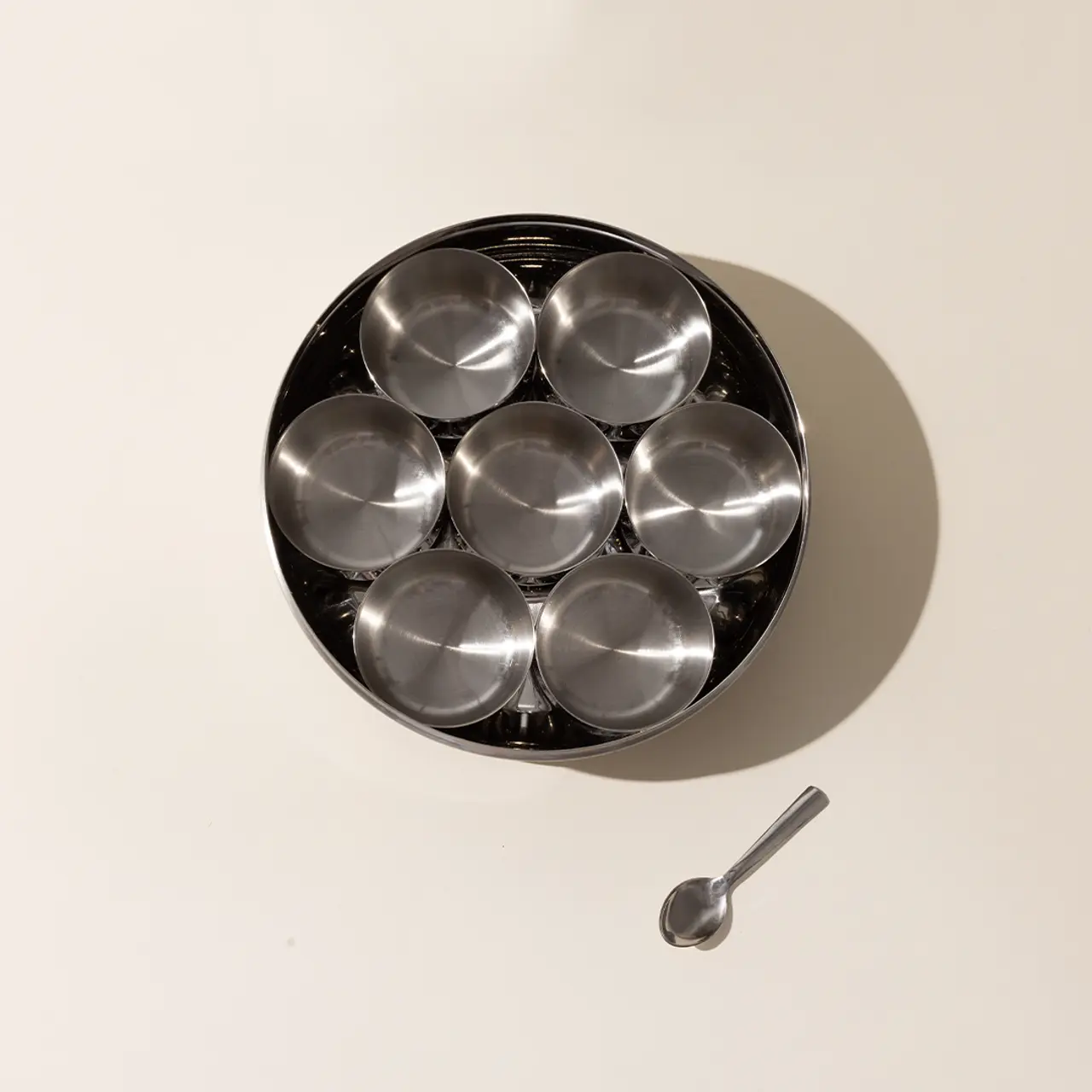 Seven empty metal bowls are neatly arranged inside a larger bowl next to a spoon on a light surface.