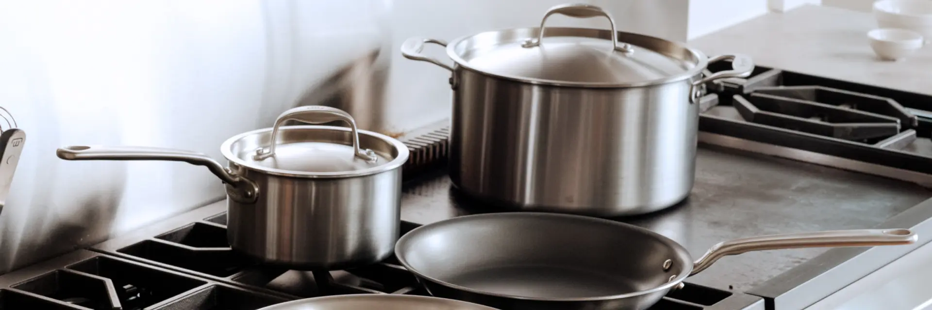 A set of stainless steel cookware, including a frying pan and two pots, sits on a modern stovetop.