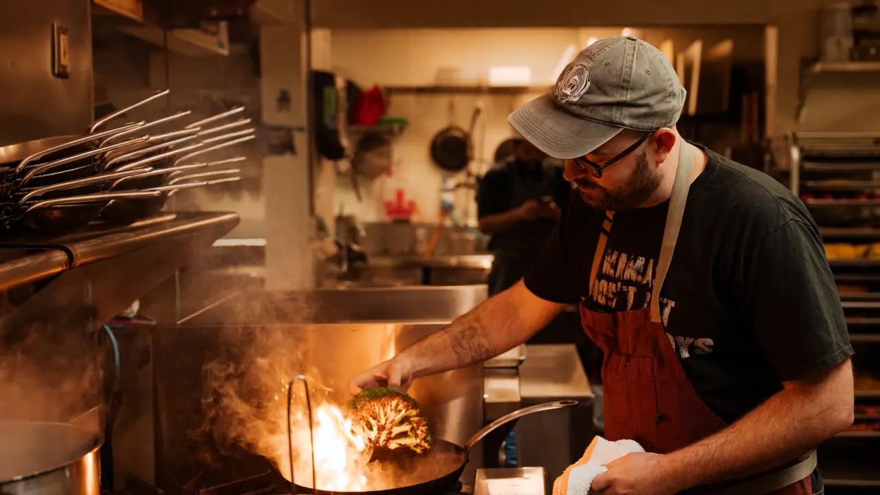 A chef attentively stirs a flaming wok amidst a bustling kitchen environment.