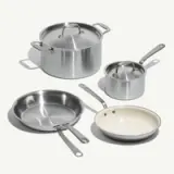 A set of four stainless steel cooking pots and pans of varying sizes, including a large pot with a lid, a medium saucepan, a large skillet, and a smaller frying pan with a white interior.