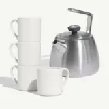 Four white coffee mugs stacked beside a sleek silver kettle.