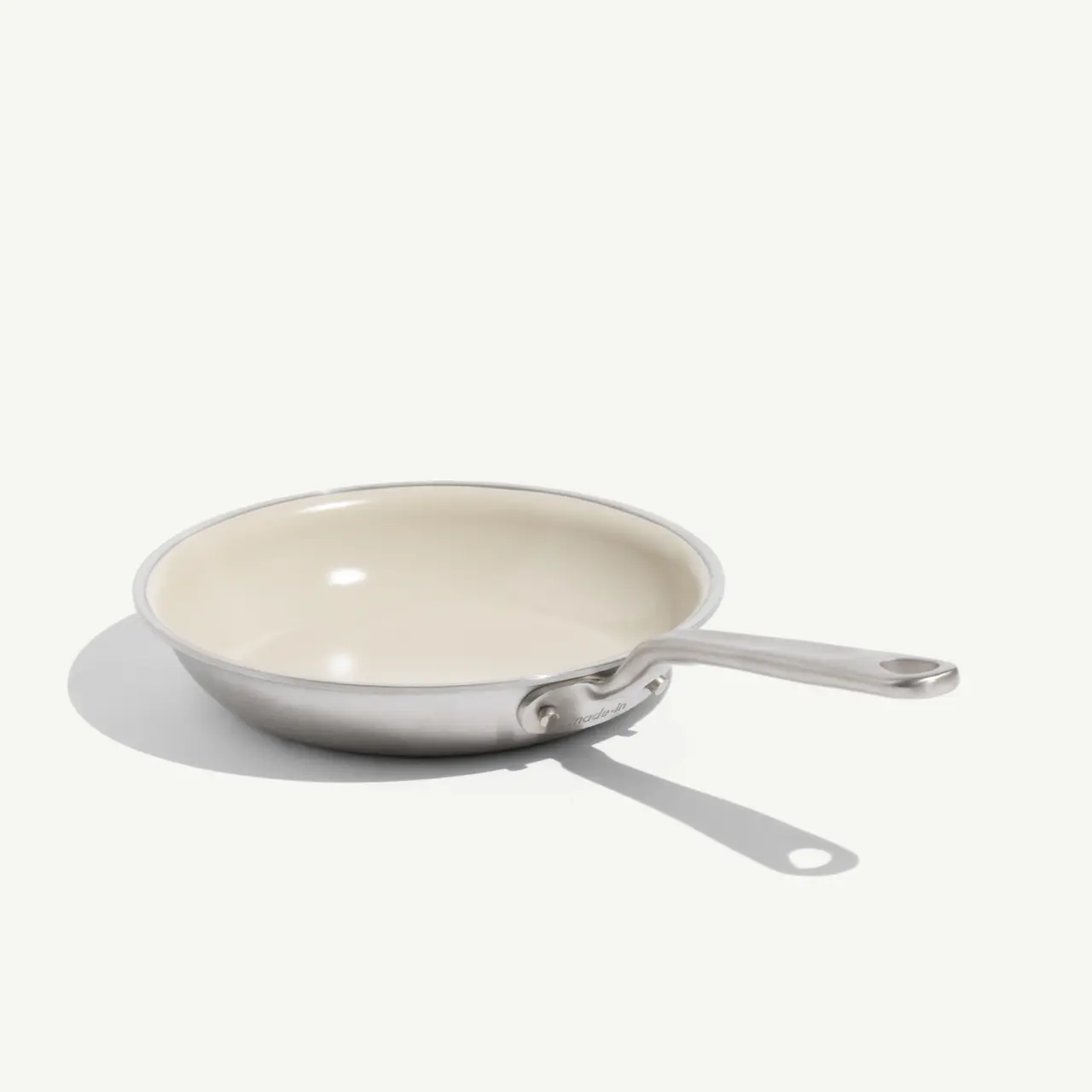 A stainless steel frying pan with a cream-colored non-stick interior and a long handle rests on a light background.