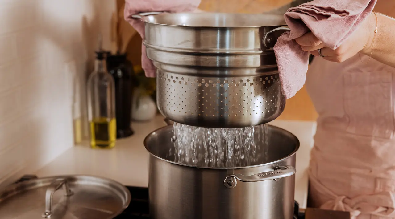 A person is draining water from a pot using a metal colander in a kitchen.