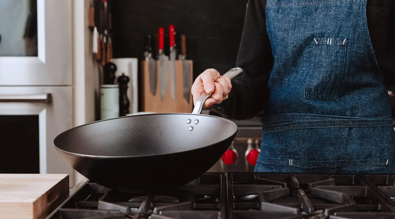 A person wearing a denim apron holds a large black frying pan over a stovetop in a kitchen.