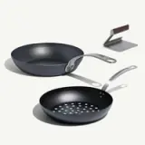 Two frying pans, one with a flat bottom and one with perforations, accompanied by a spatula, all featured in a sleek, modern design.