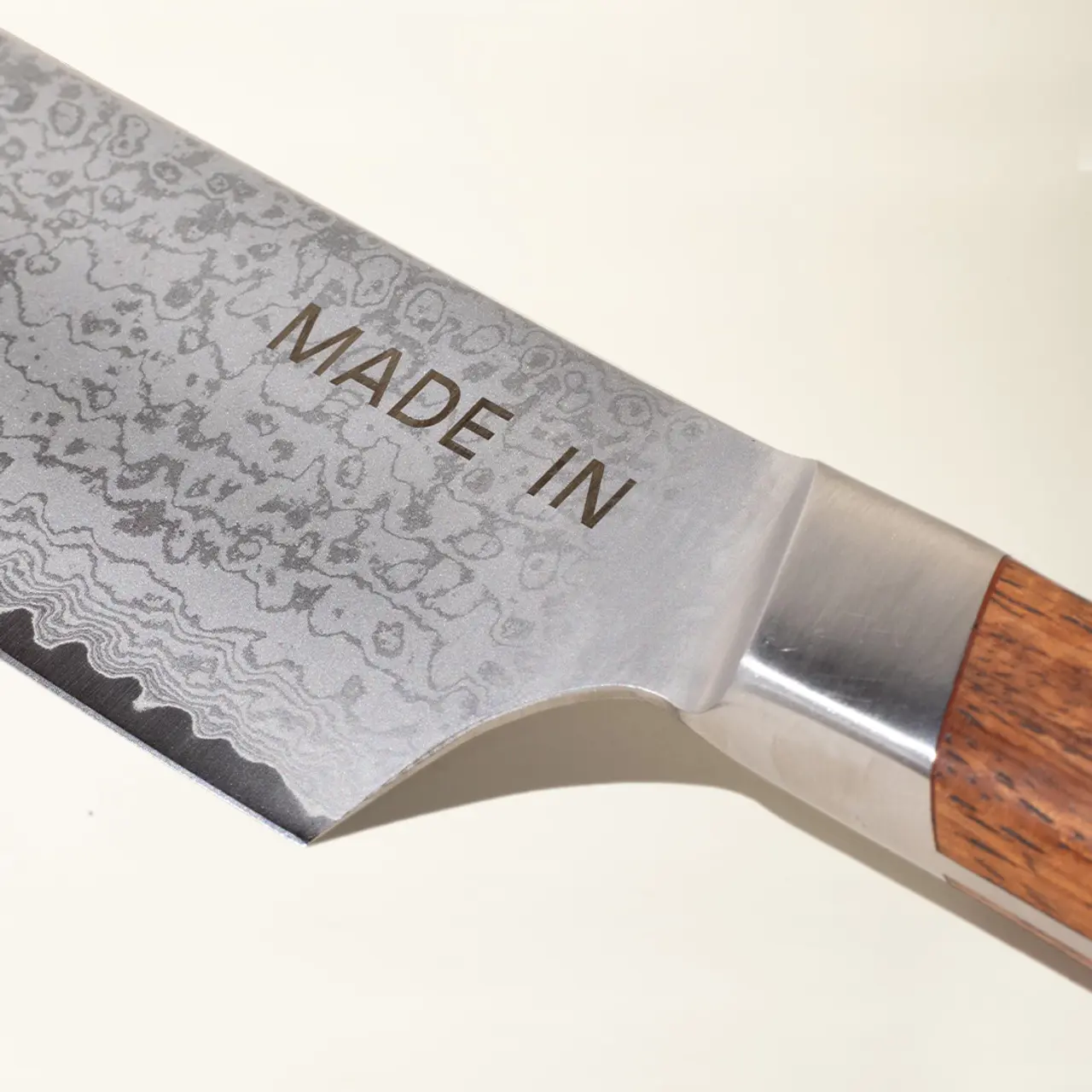 A close-up of a knife blade with a "MADE IN" inscription and a patterned, Damascus-style design near the cutting edge, transitioning to a polished steel near the bolster and a wooden handle.