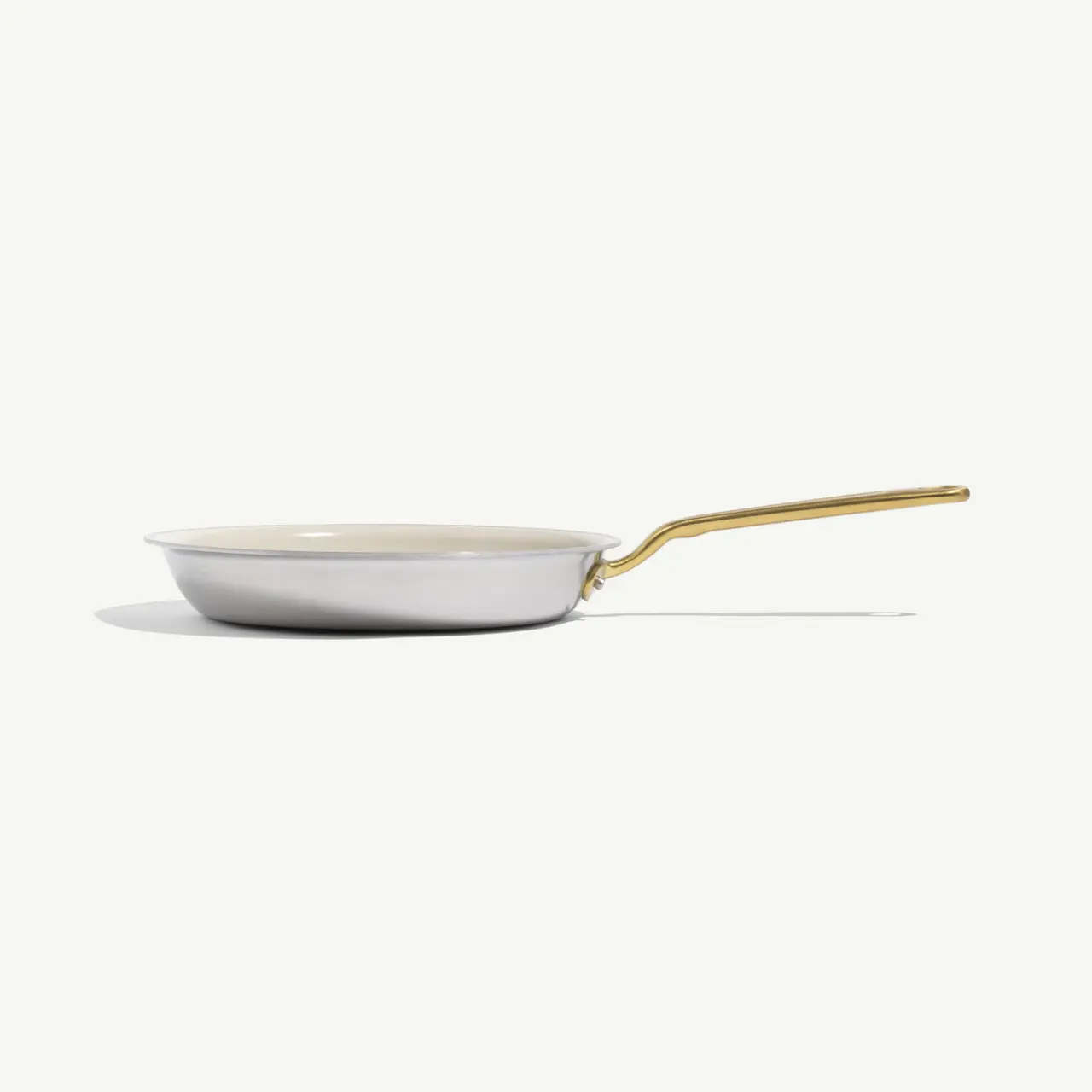 A white frying pan with a gold handle rests against a light background, casting a soft shadow.