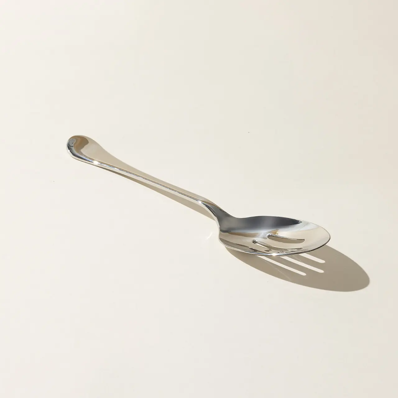 A stainless steel spork, combining features of both a spoon and a fork, casts a shadow on a light-colored surface.