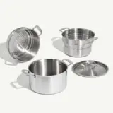 A set of stainless steel cookware including a pot, a lid, a steamer insert, and a pasta strainer is displayed.
