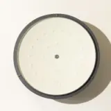 A top-down view of a large circular candle with multiple wicks, contained within a soft-edged dark container, casting subtle shadows due to a light source.