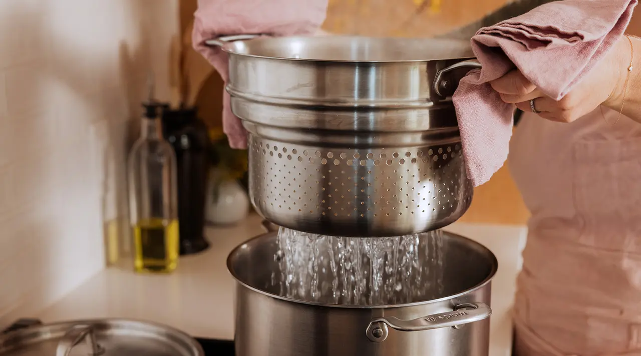 A person is draining water from a pasta pot using a colander over a kitchen sink.