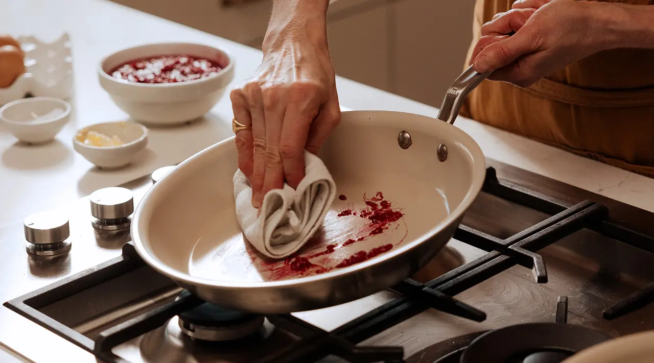 Hands are wiping a pan with a cloth on a stove top, surrounded by cooking ingredients.