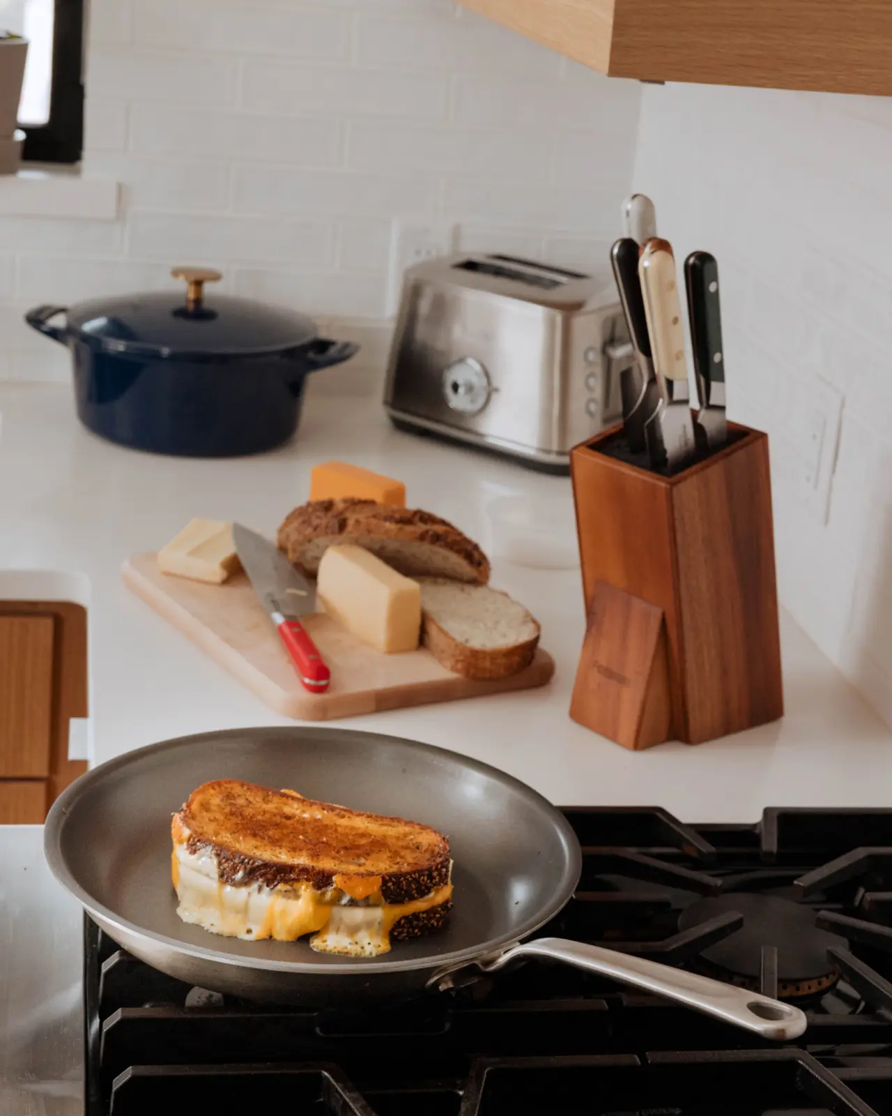 A grilled cheese sandwich is cooking in a pan on a stove, surrounded by kitchen appliances and various types of cheese on a cutting board.