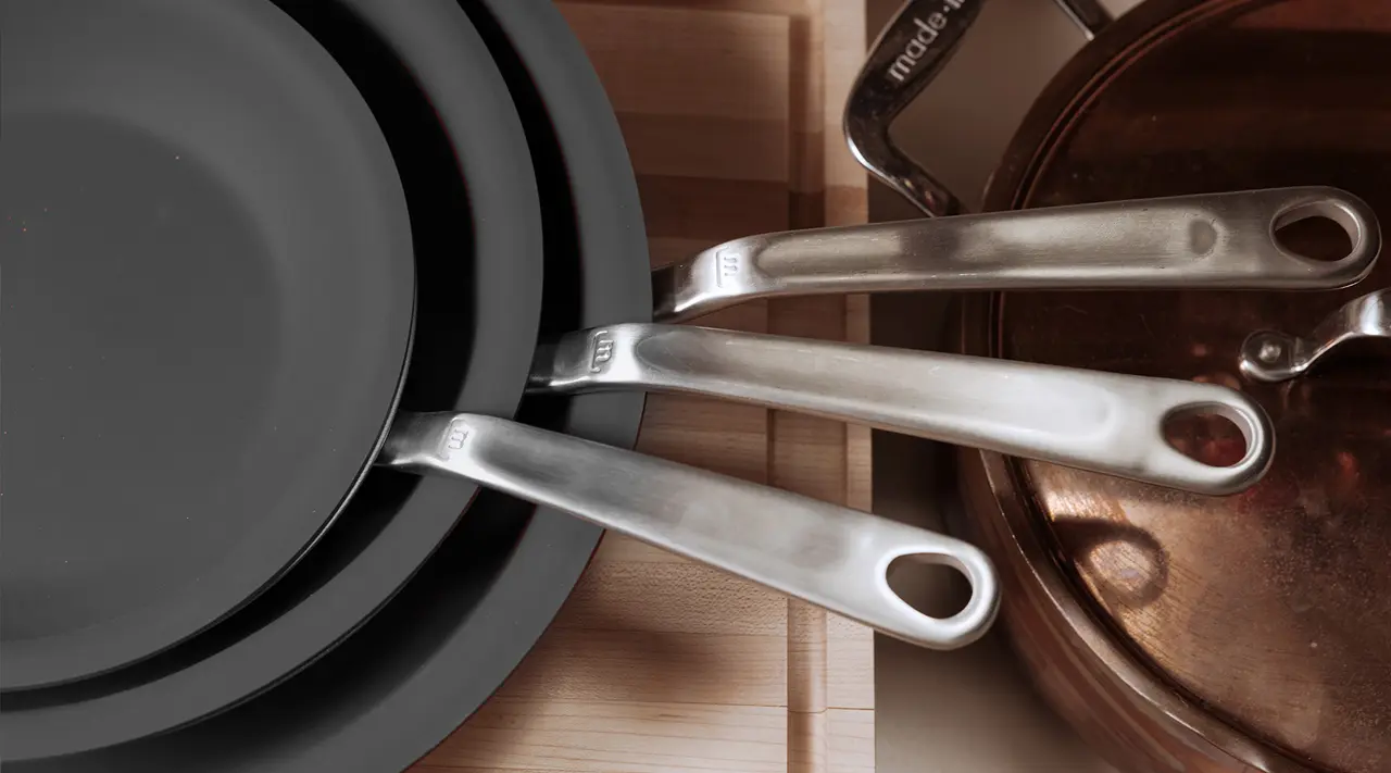 Three stacked black frying pans with shiny metal handles resting on a wooden surface, alongside a copper pot.