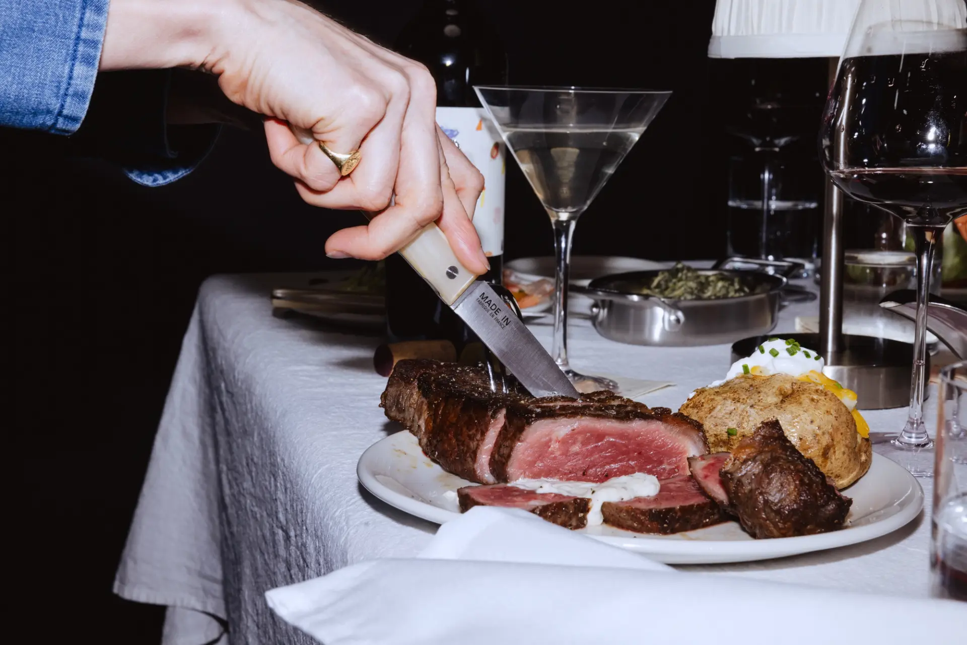 A hand is seen slicing a medium-rare steak on a plate surrounded by various side dishes and drinks.