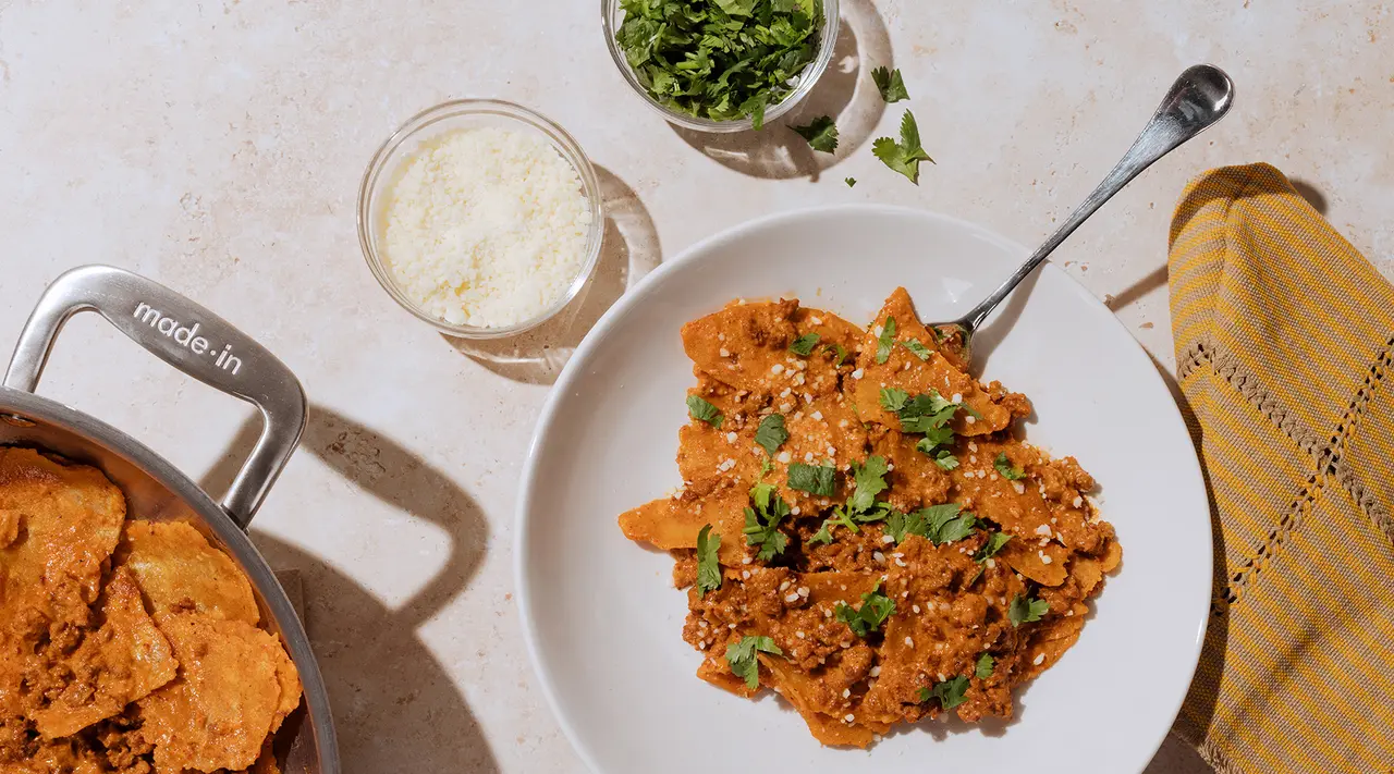 A plate of a tomato-based pasta dish garnished with herbs, next to a pot with a wooden spoon, a glass of white substance, a container of grated cheese, and a bunch of fresh herbs on a kitchen countertop.