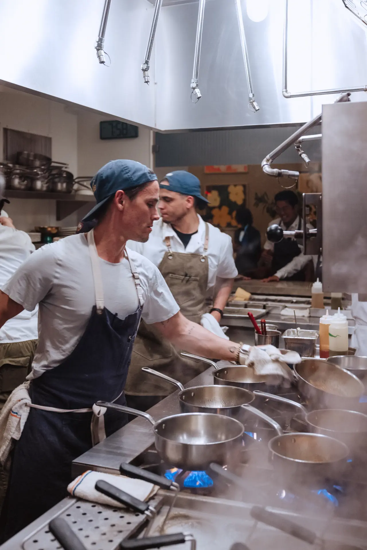 Two chefs are working in a busy kitchen, preparing dishes on the stovetop with pots steaming.