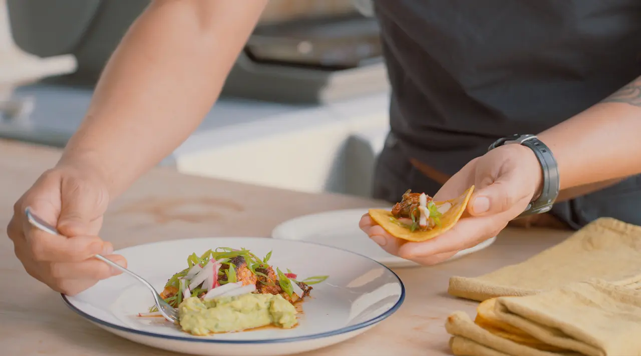 A person is plating a taco with tongs, while holding another one in their hand, on a table with a folded yellow cloth to the side.