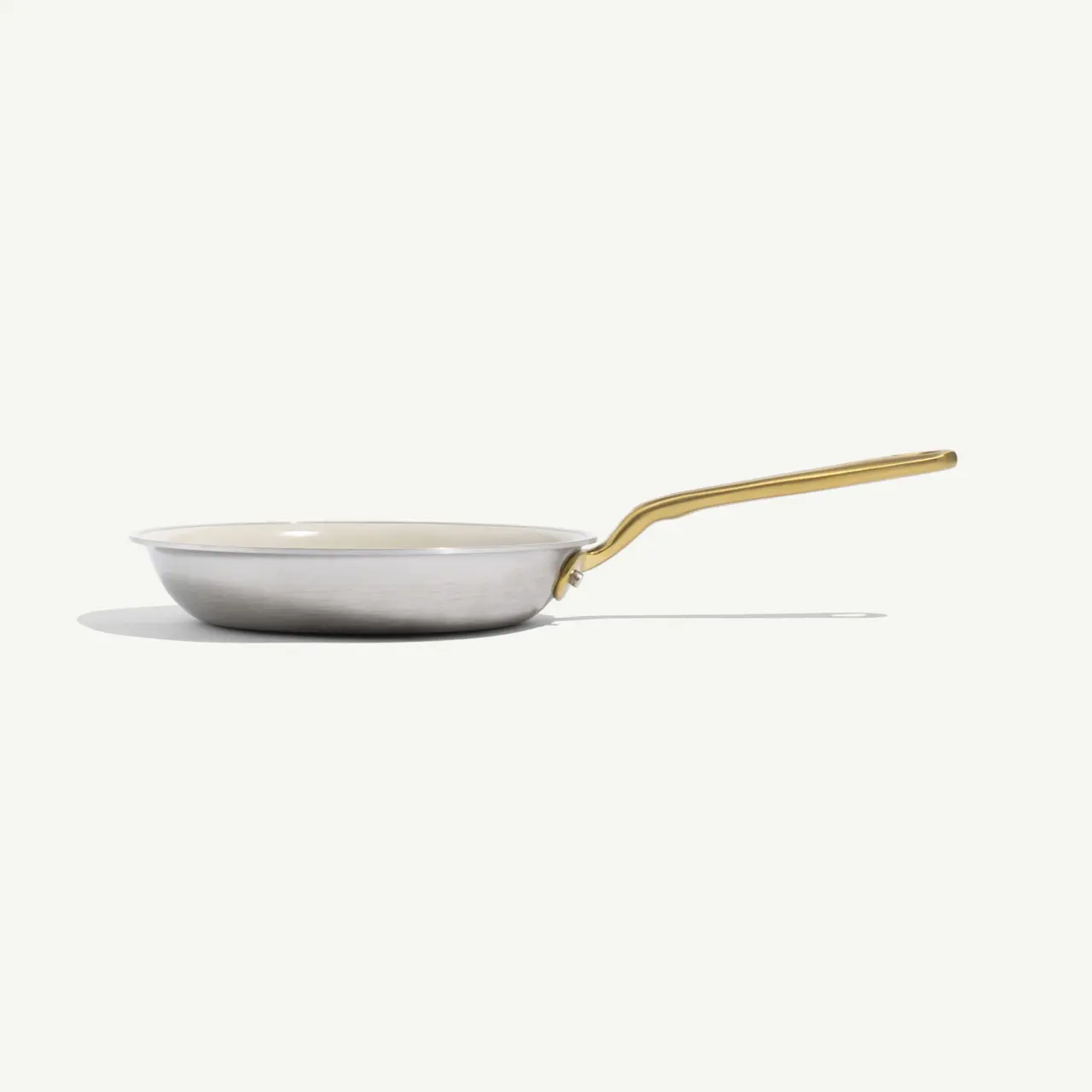 A sleek, silver frying pan with a gold handle sits against a pale background, showcasing its minimalist design.