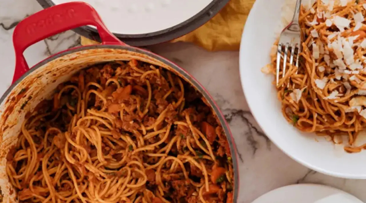 A pot of spaghetti with sauce sits next to a plate partially filled with the pasta, ready for serving.