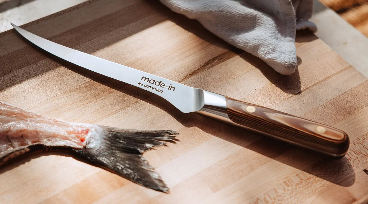 A chef's knife lies on a wooden cutting board next to a cloth and the tail of a fish under natural lighting.