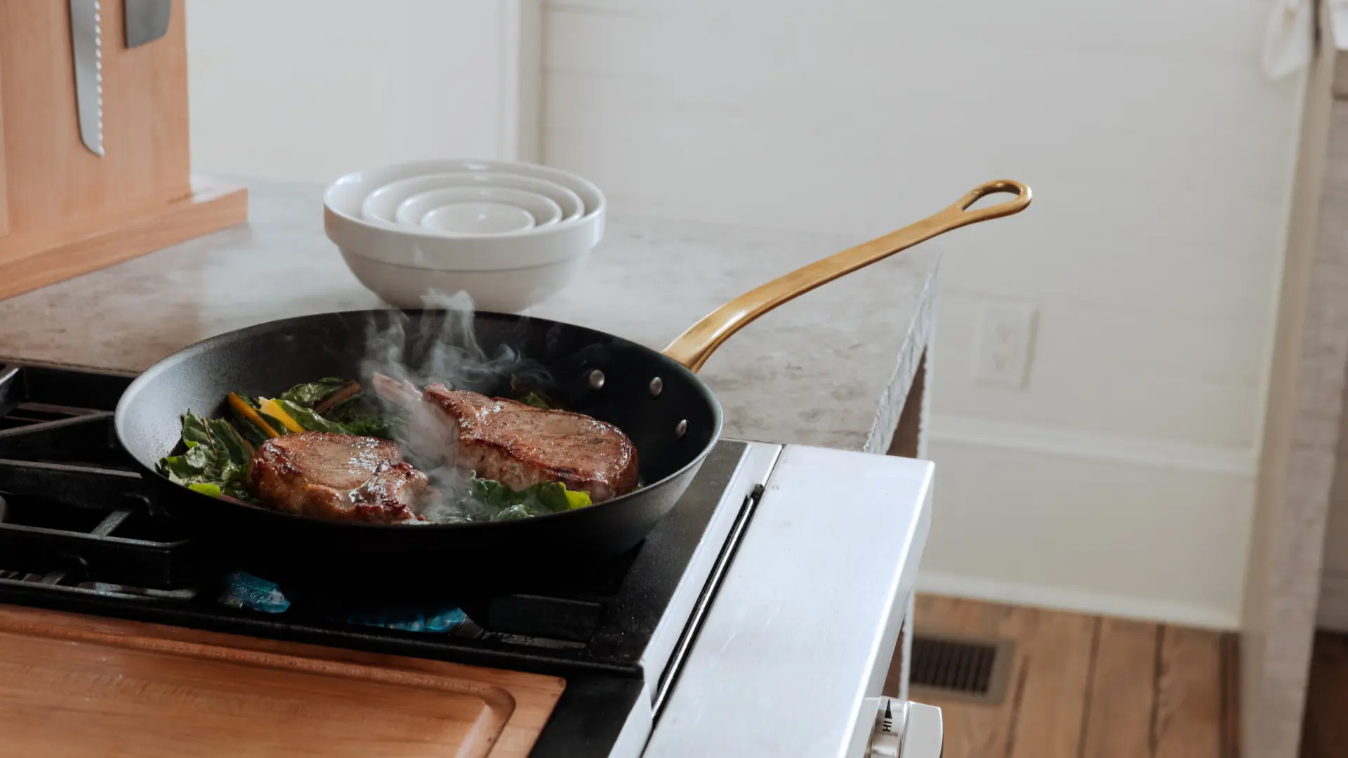 Steaks are sizzling in a frying pan on a stove with steam rising, and a bowl is seen in the background.