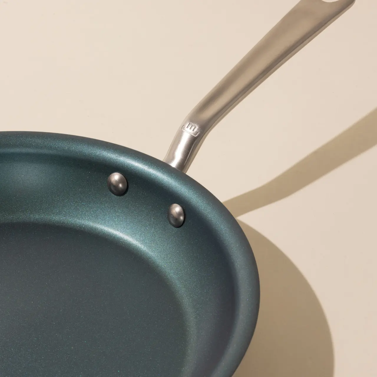 A non-stick frying pan with two metal rivets securing the handle, casting a shadow on a light background.